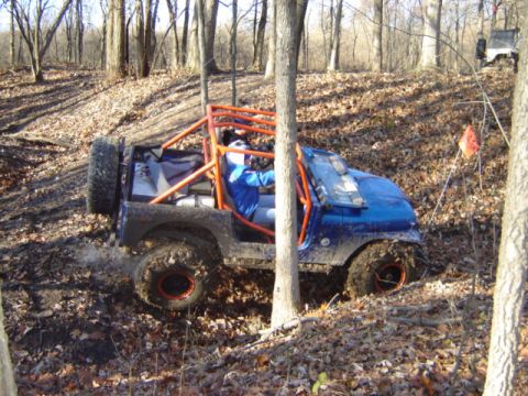 1992 Ford Bronco - A blue off-road buggy navigates a muddy trail in a wooded area, with another vehicle in the distance.