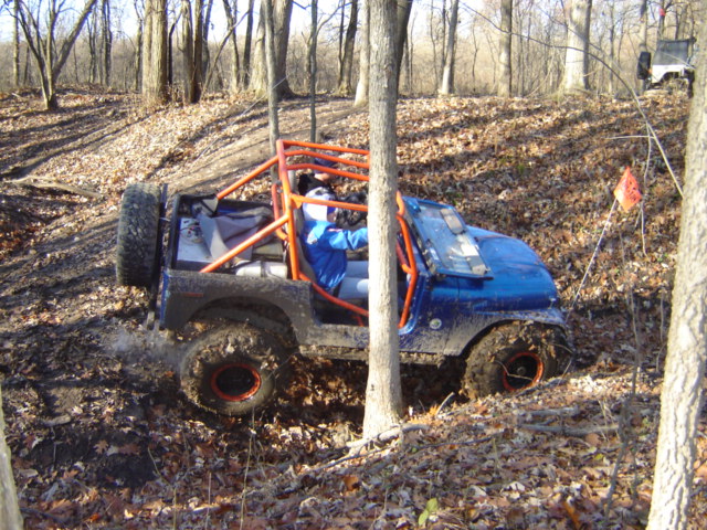 Blue buggy with orange roll cage driving through muddy woods. Another white vehicle is visible on a hill in the background