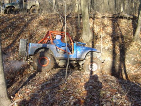 1992 Ford Bronco - Two off-road vehicles navigate a muddy, leaf-covered hill in a wooded area.