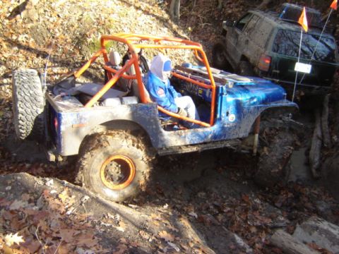 1992 Ford Bronco - Two off-road vehicles navigate a muddy, wooded trail with fallen leaves.