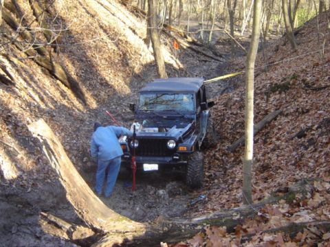 1992 Ford Bronco - A person attempts to free a black Jeep Wrangler from a muddy ditch in a wooded area.