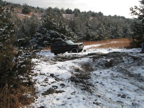 1995 Ford Bronco - A Jeep Cherokee XJ navigates a snowy and muddy path surrounded by pine trees.