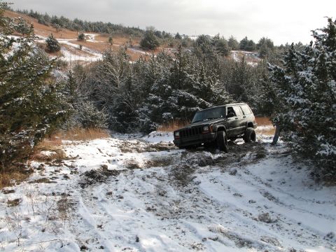 1995 Ford Bronco - A dark green Jeep Cherokee XJ navigates a challenging snowy and muddy trail through a wooded landscape.