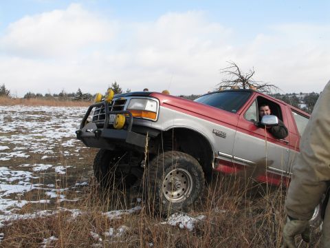1995 Ford Bronco - A red and silver Ford Bronco is driven off-road through a field with sparse snow cover.