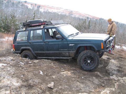 1995 Ford Bronco - A modified Jeep Cherokee XJ is stuck in the mud on a rocky incline, with a person standing nearby.