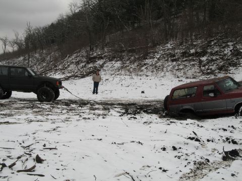 1995 Ford Bronco - A red and gray Ford Bronco is stuck in the mud, being pulled by a black Jeep in a snowy landscape.