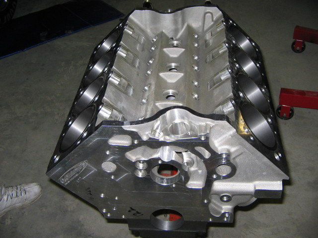Bare aluminum V8 engine block standing upright on shop floor, showing cylinder bores and front face.