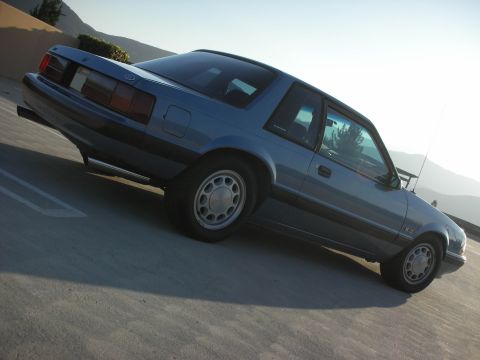 1990 Ford Mustang - Blue Fox-body Ford Mustang LX 5.0 notchback coupe parked, rear three-quarter view with mountains behind.