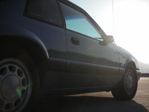 1990 Ford Mustang - A 1990 Ford Mustang coupe is seen from the side under bright sunlight.