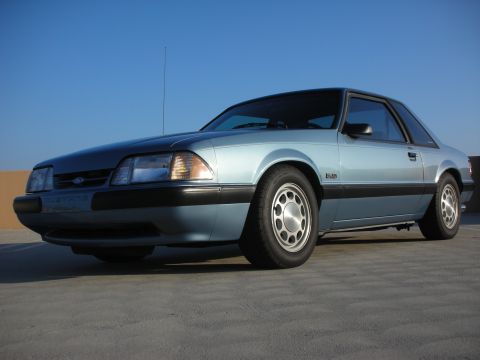 1990 Ford Mustang - A light blue 1990 Ford Mustang coupe is shown from a 3/4 front angle against a clear blue sky.