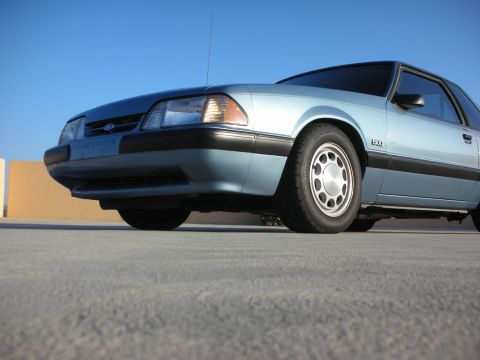 1990 Ford Mustang - A light blue 1990 Ford Mustang 5.0 coupe is shown from a low angle, highlighting its front quarter view against a clear blue sky.
