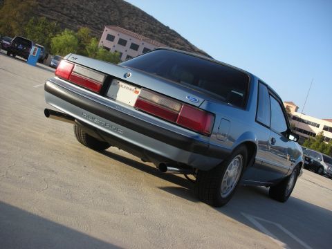 1990 Ford Mustang - The rear of a blue 1990 Ford Mustang is shown parked in a lot with buildings and hills in the background.