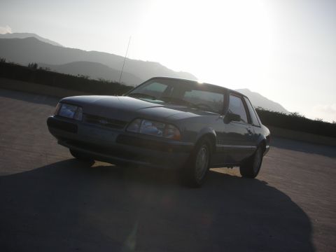 1990 Ford Mustang - A 1990 Ford Mustang coupe is viewed from a 3/4 front angle against a backdrop of hazy mountains at sunset.