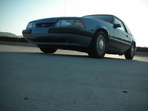 1990 Ford Mustang - A blue 1990 Ford Mustang 5.0 coupe is shown from a low angle, highlighting its front three-quarter view.