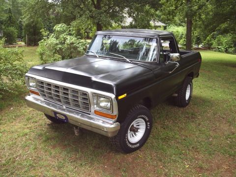 1978 Ford Bronco - Black late-1970s Ford Bronco parked on grass in a shaded yard, shot from the driver's-side front three-quarter with a flat-black hood, silv…