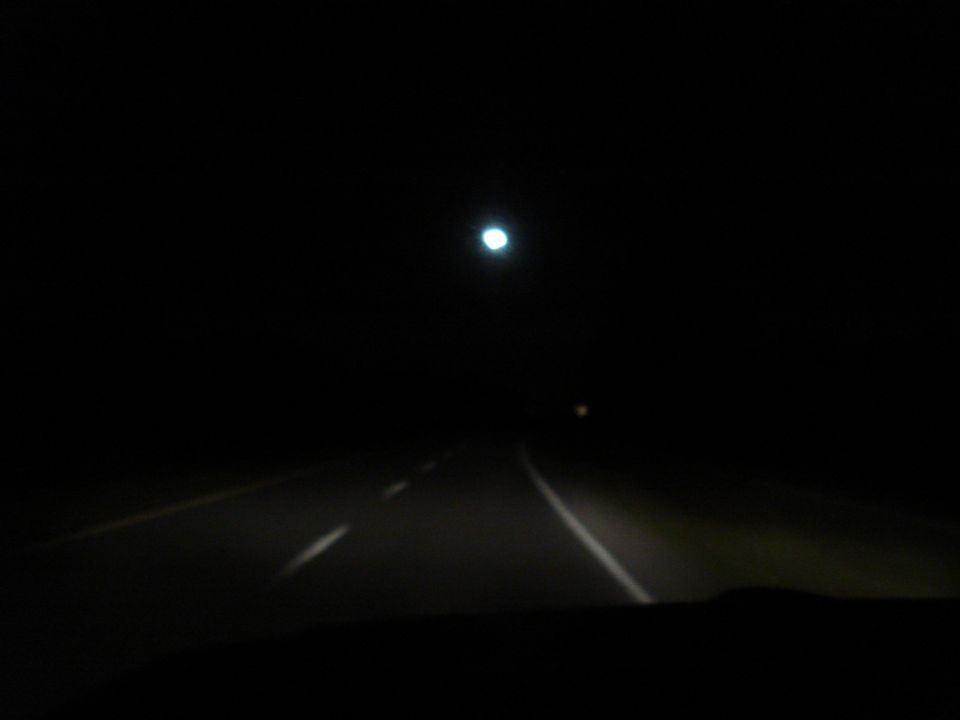 View from inside a car driving on a dark road at night, with headlights illuminating the asphalt and a distant light source overhead