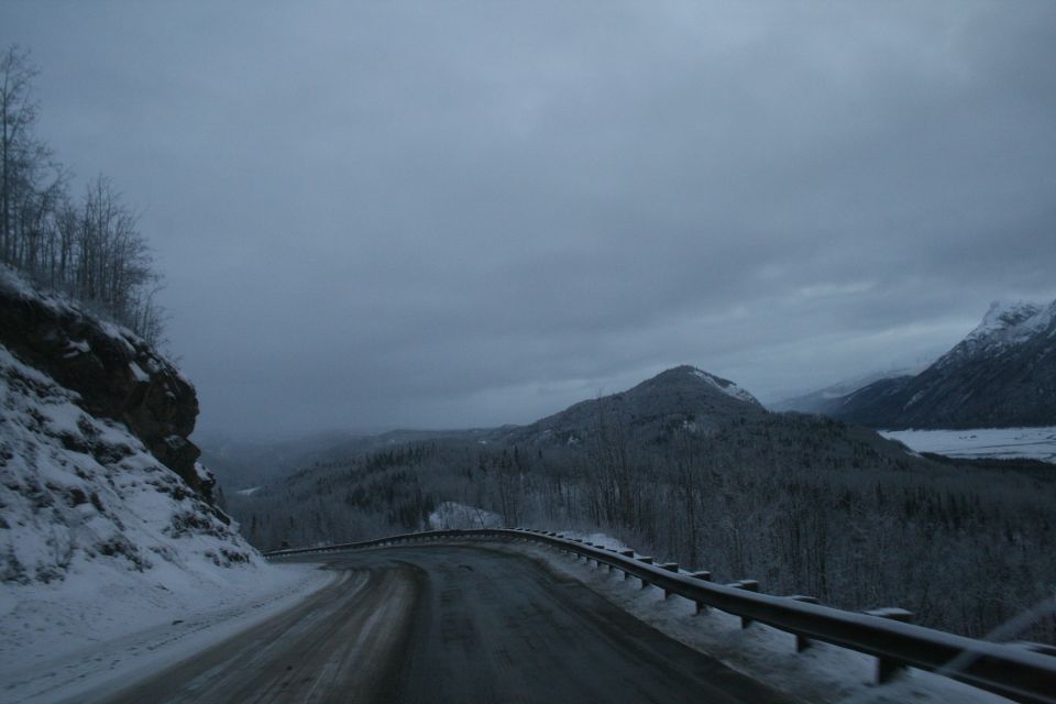 Winding mountain road covered in snow with trees and mountains in the background under a cloudy sky