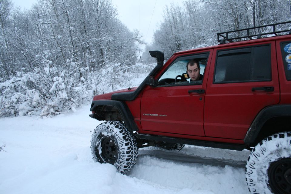 Red Jeep Cherokee XJ with snorkel and roof rack driving in snow. Driver looks at camera