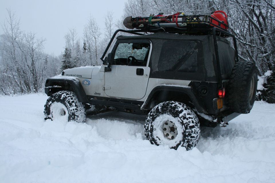 White Jeep Wrangler in deep snow, roof rack with shovel and red gas can, Rubicon badge visible