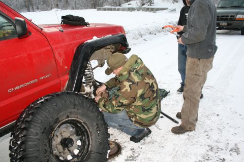 2003 Jeep TJ - A red Jeep Cherokee Sport is being worked on by a person in camouflage, with another vehicle visible in the background during a snowy day.
