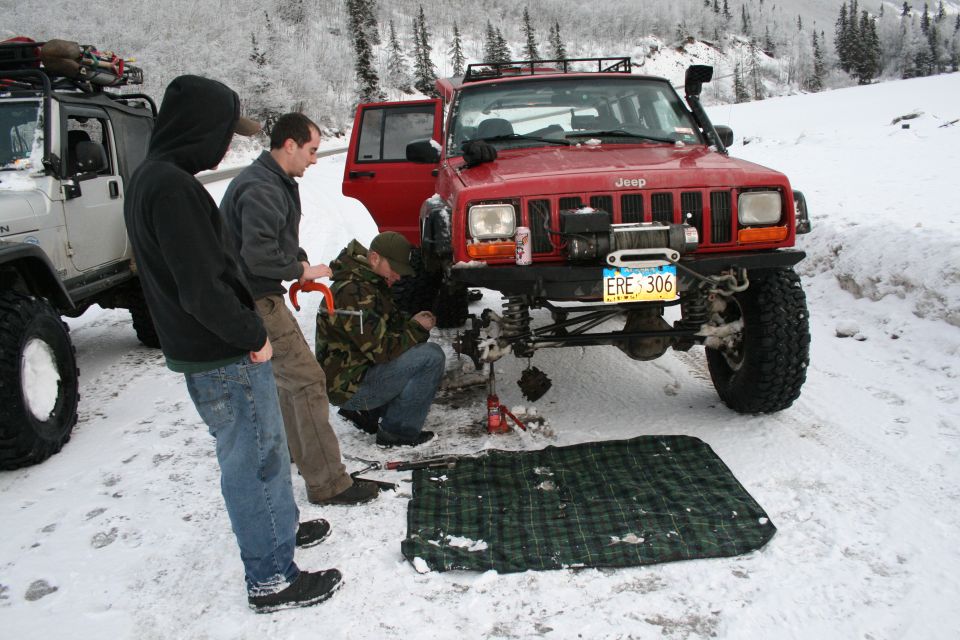 Red Jeep Cherokee XJ with winch and aftermarket bumper, being worked on by three people in the snow. A white Jeep Wrangler is parked to the left