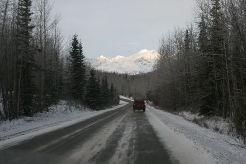 2003 Jeep TJ - A red SUV travels on a winter road surrounded by trees, with snowy mountains ahead.