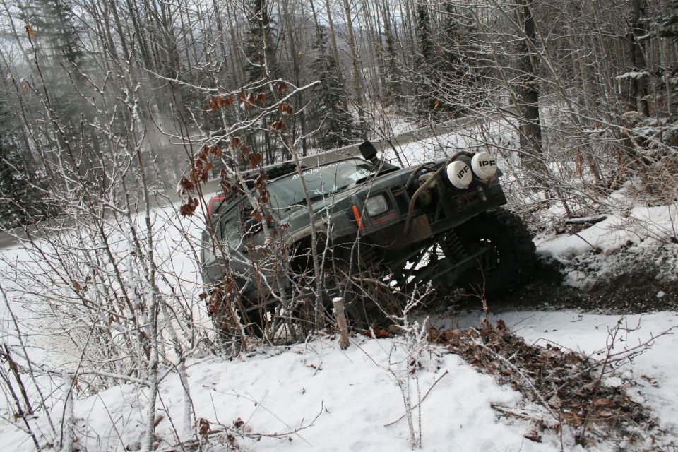 Modified Jeep Cherokee XJ with large tires and auxiliary lights driving off-road in a snowy forest