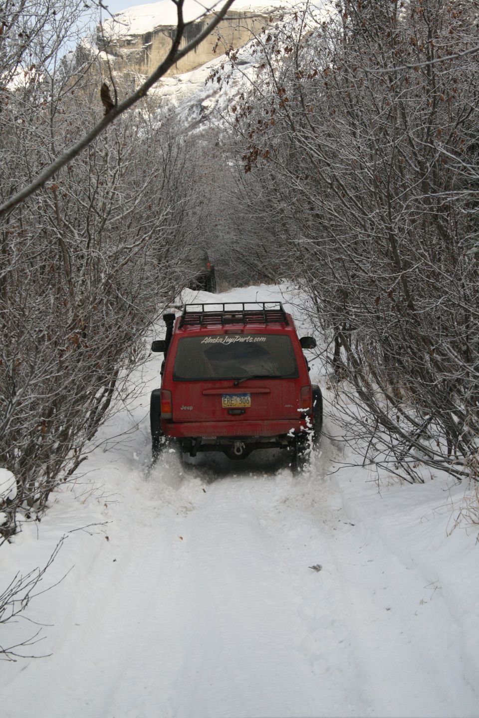 Red Jeep Cherokee XJ driving on a snow-covered trail, kicking up snow. A second vehicle is visible further down the path