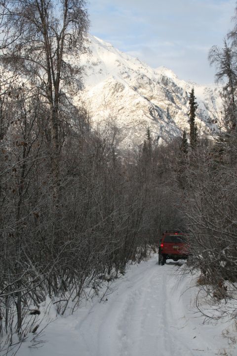 2003 Jeep TJ - A red SUV navigates a snowy path surrounded by bare trees, with a snow-covered mountain range looming ahead.