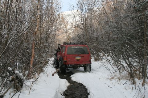 2003 Jeep TJ - Red Jeep Cherokee XJ navigates a snowy, wooded path. The vehicle has a roof rack and a sticker on the rear window.