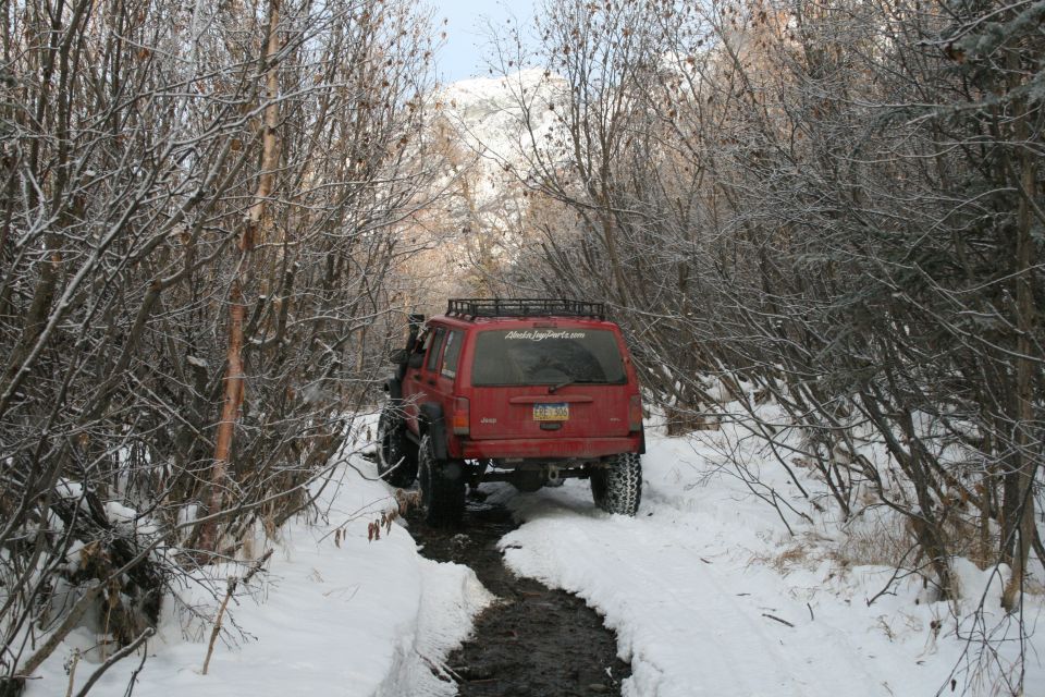 Red Jeep Cherokee XJ with a roof rack driving on a muddy, snowy trail between bare trees. License plate is visible