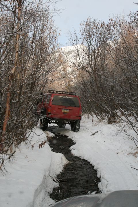 2003 Jeep TJ - A red Jeep Cherokee XJ navigates a challenging snowy and muddy path surrounded by bare trees.