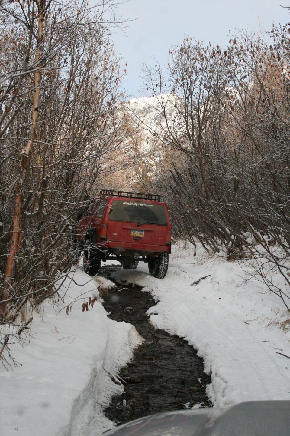 Red Jeep Cherokee XJ driving through a snowy, muddy trail. Another vehicle is partially visible in the foreground