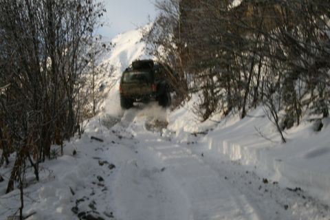 2003 Jeep TJ - A dark SUV navigates a snow-covered mountain path, kicking up snow as it drives.