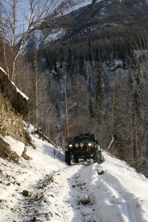 2003 Jeep TJ - A black SUV navigates a snow-covered mountain path with dense trees and a rocky incline.