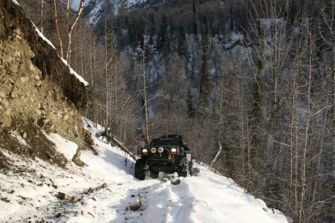 2003 Jeep TJ - A black off-road vehicle navigates a snowy mountain path surrounded by bare trees and a steep embankment.