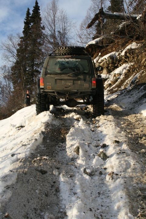 2003 Jeep TJ - A Jeep Cherokee XJ navigates a challenging snowy and muddy incline, with a spare tire mounted on its roof rack.