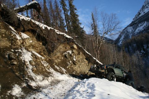 2003 Jeep TJ - A person pushes a green off-road vehicle up a steep, snowy mountain path with a landslide visible on the left.
