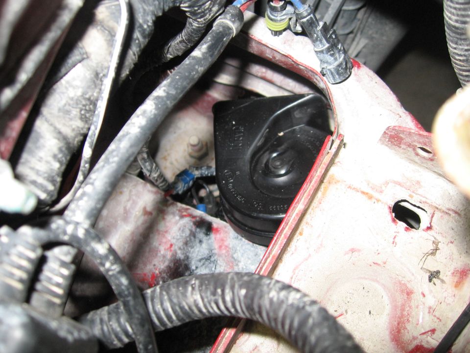 Close-up of a black vehicle horn and surrounding wiring harness, showing red painted metal structure