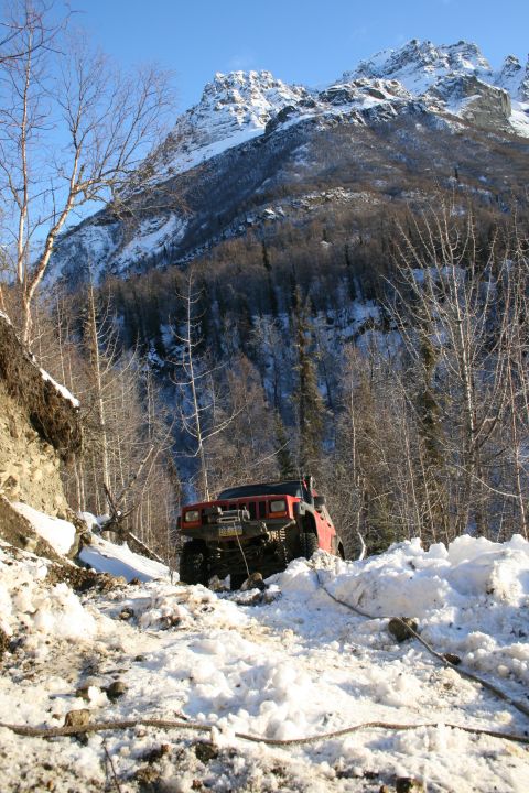 2003 Jeep TJ - A red Jeep Cherokee navigates a challenging snowy path on a mountain trail, with a winch cable extending from the front.