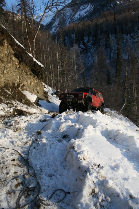 2003 Jeep TJ - A red Jeep Cherokee XJ navigates a snow-covered trail in a mountainous, wooded area.