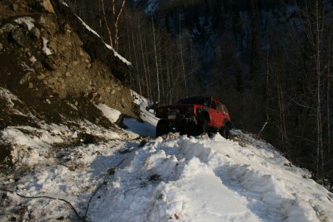 2003 Jeep TJ - A red Jeep Cherokee XJ navigates a treacherous snowy incline on a wooded mountainside.