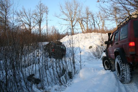 2003 Jeep TJ - A red Jeep Cherokee navigates a snowy trail while another Jeep appears stuck in the ditch beside the path.