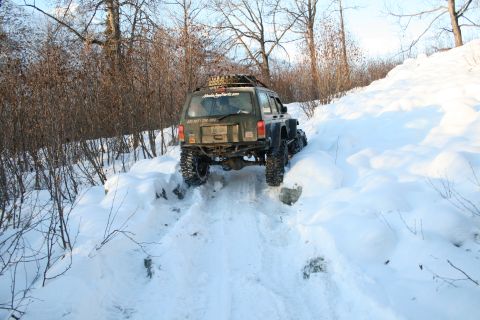 2003 Jeep TJ - Modified Jeep Cherokee XJ navigates a steep, snow-covered trail. The vehicle has large off-road tires and a roof rack.