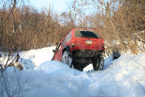 2003 Jeep TJ - A red Jeep Cherokee XJ navigates a snowy trail, its rear end visible as it climbs the incline.