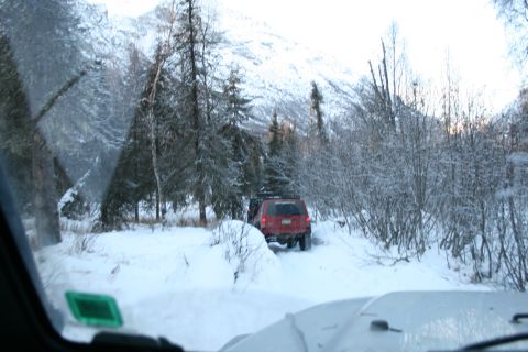 2003 Jeep TJ - A red SUV navigates a snowy path through a forest, with a mountain range visible ahead.