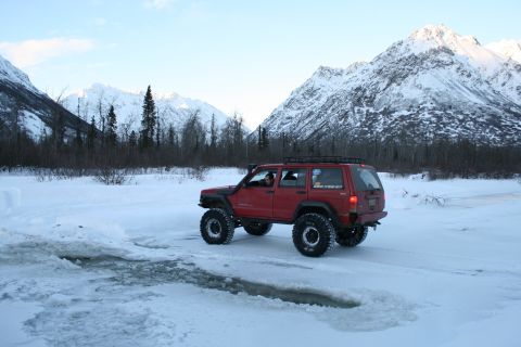2003 Jeep TJ - A red Jeep Cherokee XJ navigates a snowy landscape with mountains in the background.