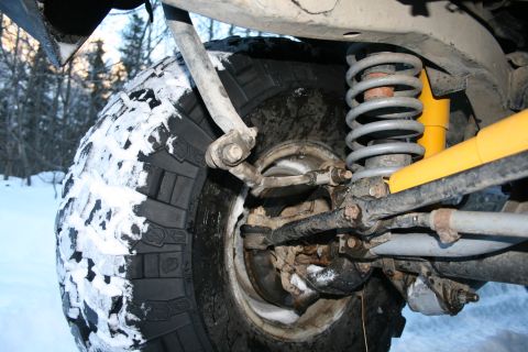 2003 Jeep TJ - The front suspension and tire of a Jeep are visible, partially covered in snow, with trees in the background.