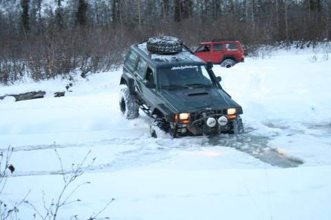2003 Jeep TJ - A dark green Jeep Cherokee XJ navigates a snowy path, with a red Jeep visible in the background.