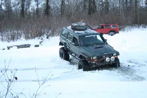 2003 Jeep TJ - Two modified Jeeps navigate a snowy, wooded area with one vehicle partially submerged in slush.